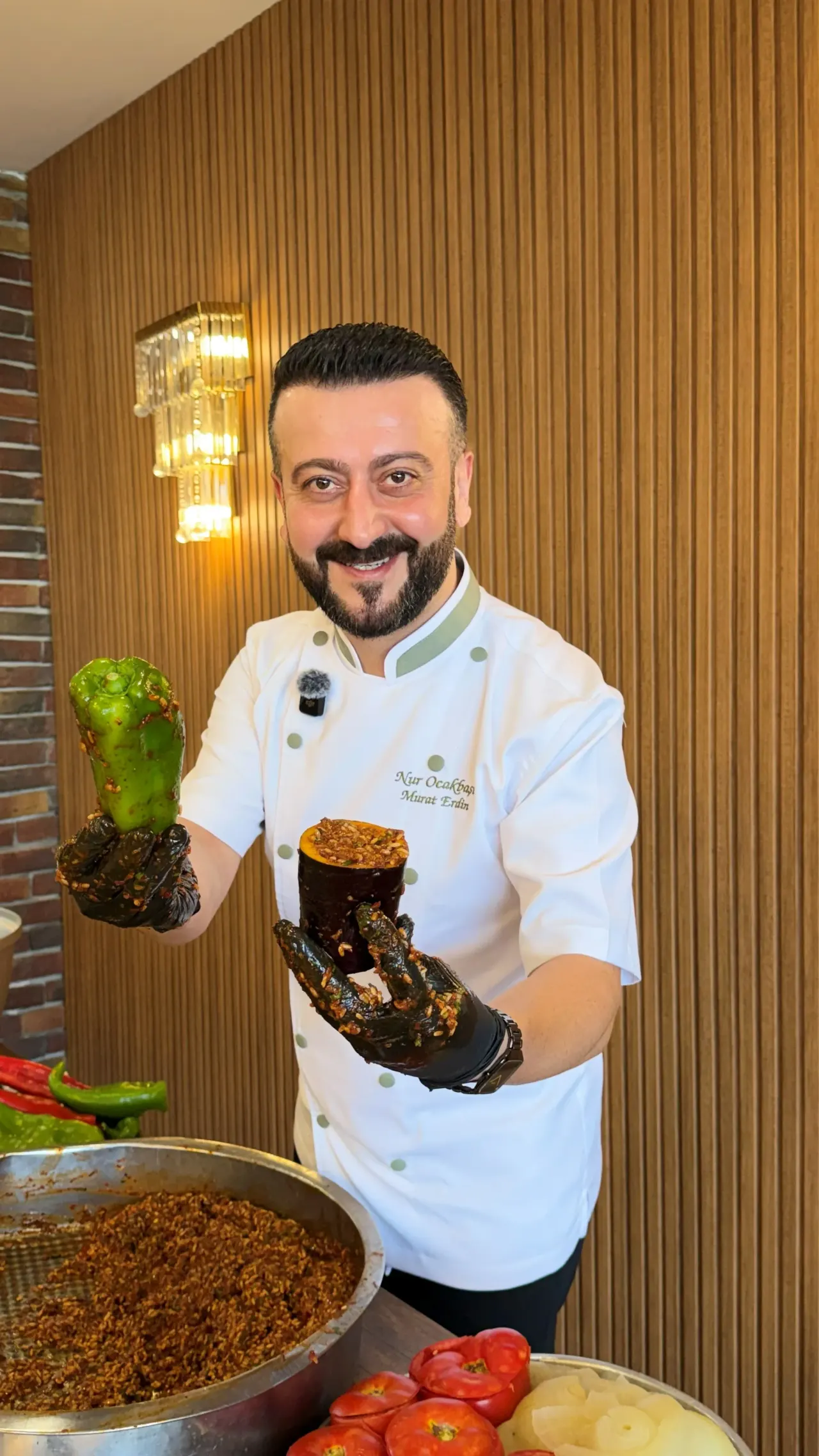 A cook holding a green pepper in one hand and an aubergine in the other, with a large pot of rice in front of him