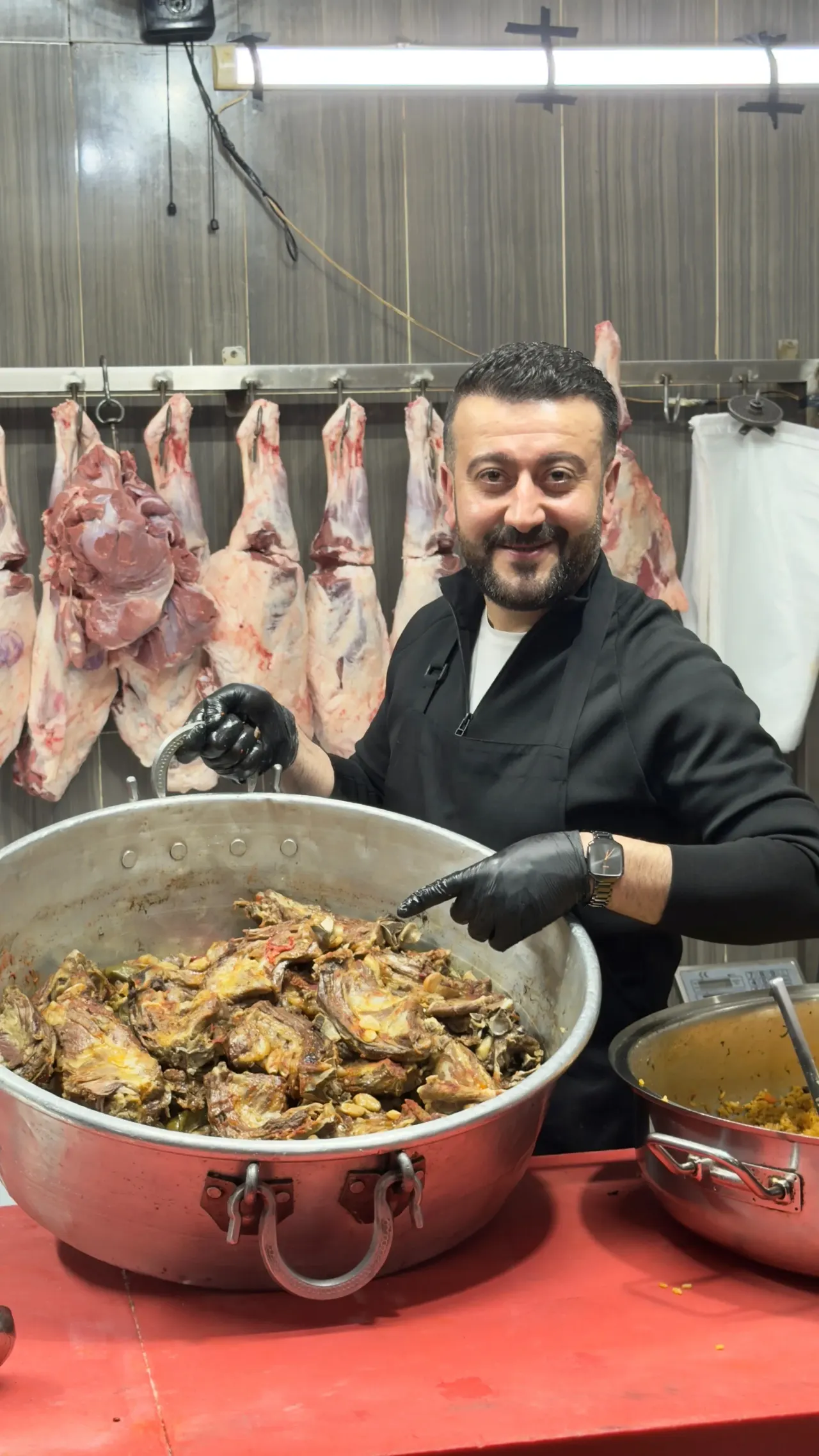 A cook in a black shirt at a large pot of cooked meat, with the butcher’s counter behind